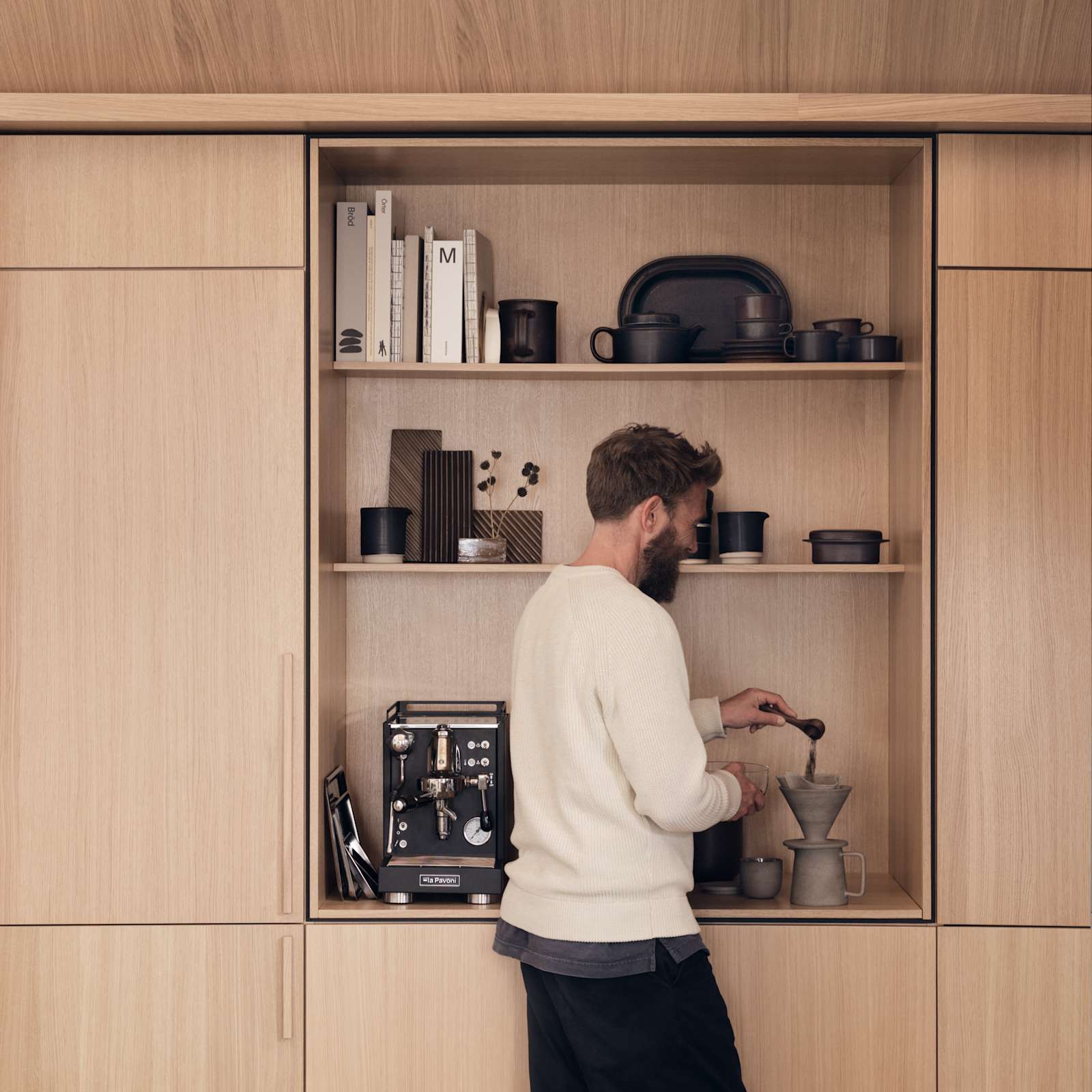 En mand måler kaffe op i Forever Oak Natur Eg-køkkenet fra INVITA i egetræsfiner med lodret træåring. Højskabe og et åbent skab med plads til kaffemaskine, indrammet med en skygge, der skaber en arkitektonisk premium følelse.