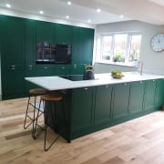 View of kitchen island and matching tall cabinets in Ashleigh's modern vibrant green Shaker Dunham, with marbled worktop and brass details like tap and cup handles.