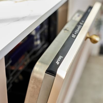 Close-up view of slightly open integrated Smeg dishwasher in Ludlow Chalk Blush cabinetry from Magnet, with a brass handle and Dekton worktop.