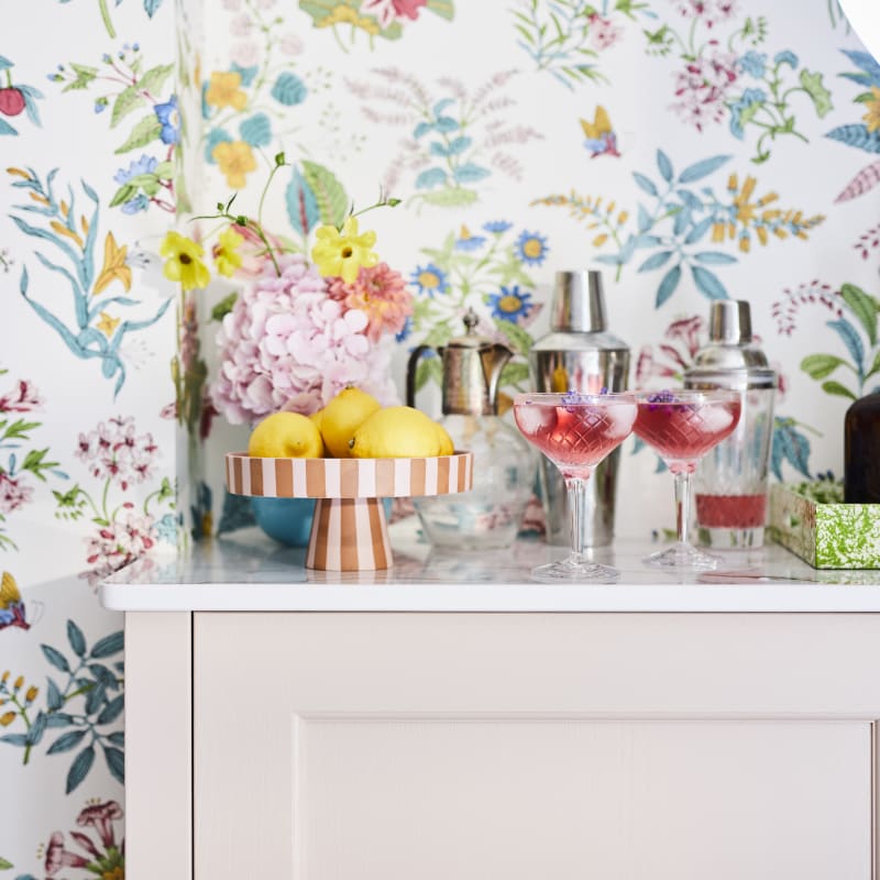 Cocktail corner in Sophie Robinsons maximalist Ludlow kitchen from Magnet, with two pink drinks poured in glasses, floral pattern wallpaper.