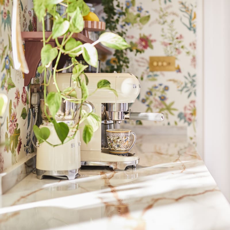 Side view of Smeg coffee machines on countertop in the coffee area in Sophie Robinson's maximalist kitchen, a plant on open shelving, a floral wallpaper.