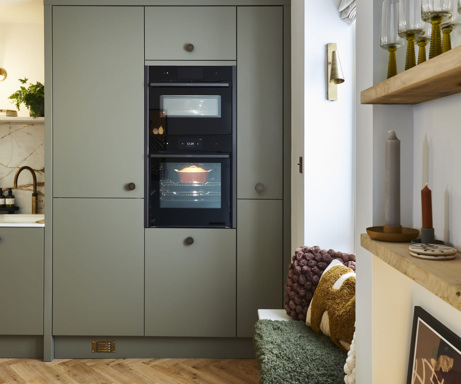 Soho Balmoral tall cabinets with integrated ovens and a bench in the window, next to a wall of open wood shelves decorated with plants, candles and wine glasses.