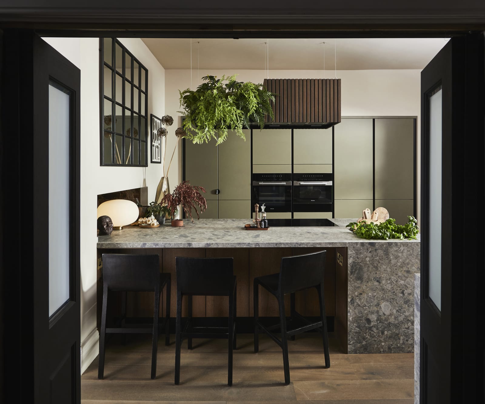 View through opened dark wood doors of the biophilic Dramatic neutral Balmoral Soho and Nordic Nature kitchen from Magnet, matching dark wood chairs by kitchen island bar seating area.