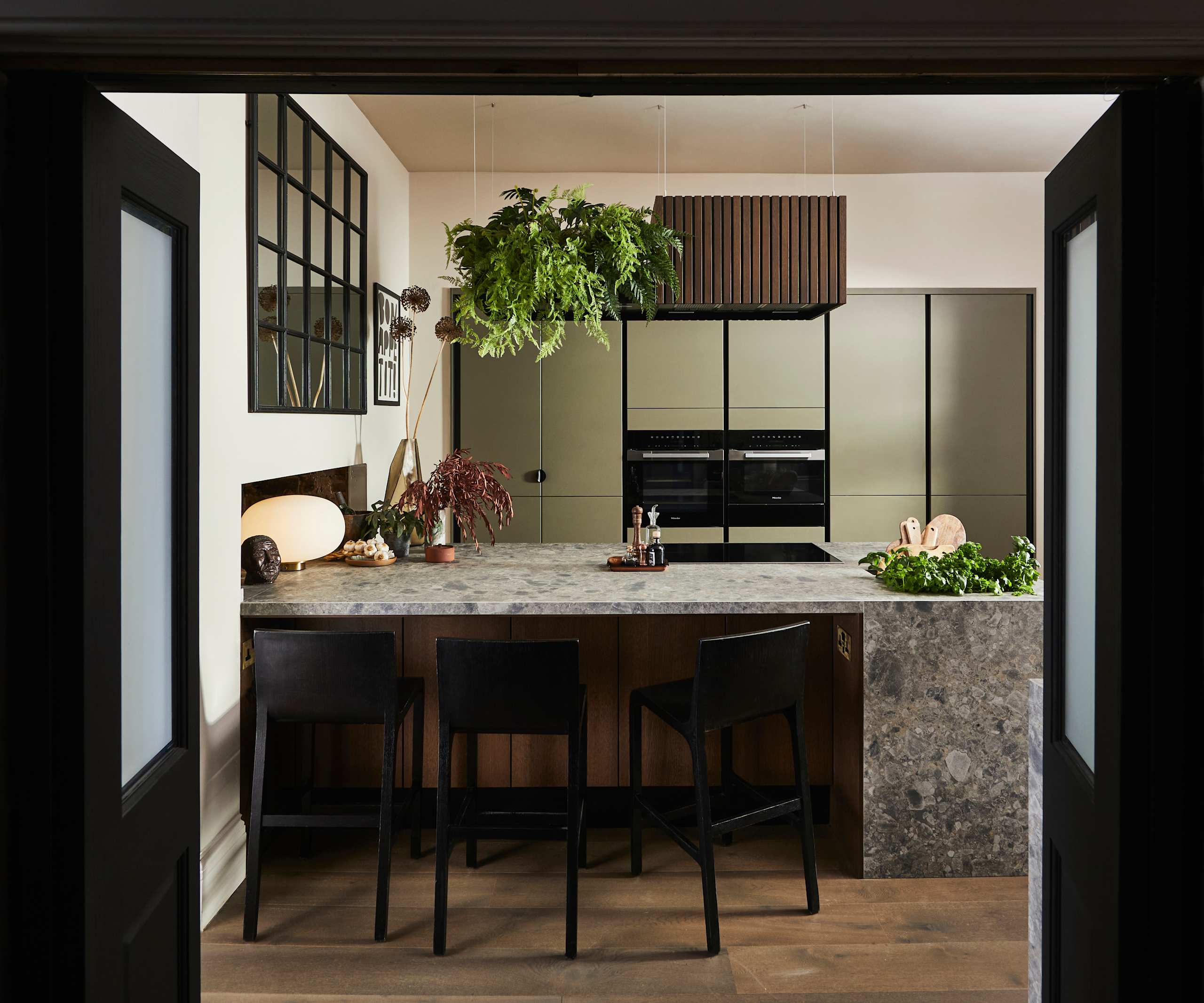 View through opened dark wood doors of the biophilic Dramatic neutral Balmoral Soho and Nordic Nature kitchen from Magnet, matching dark wood chairs by kitchen island bar seating area.