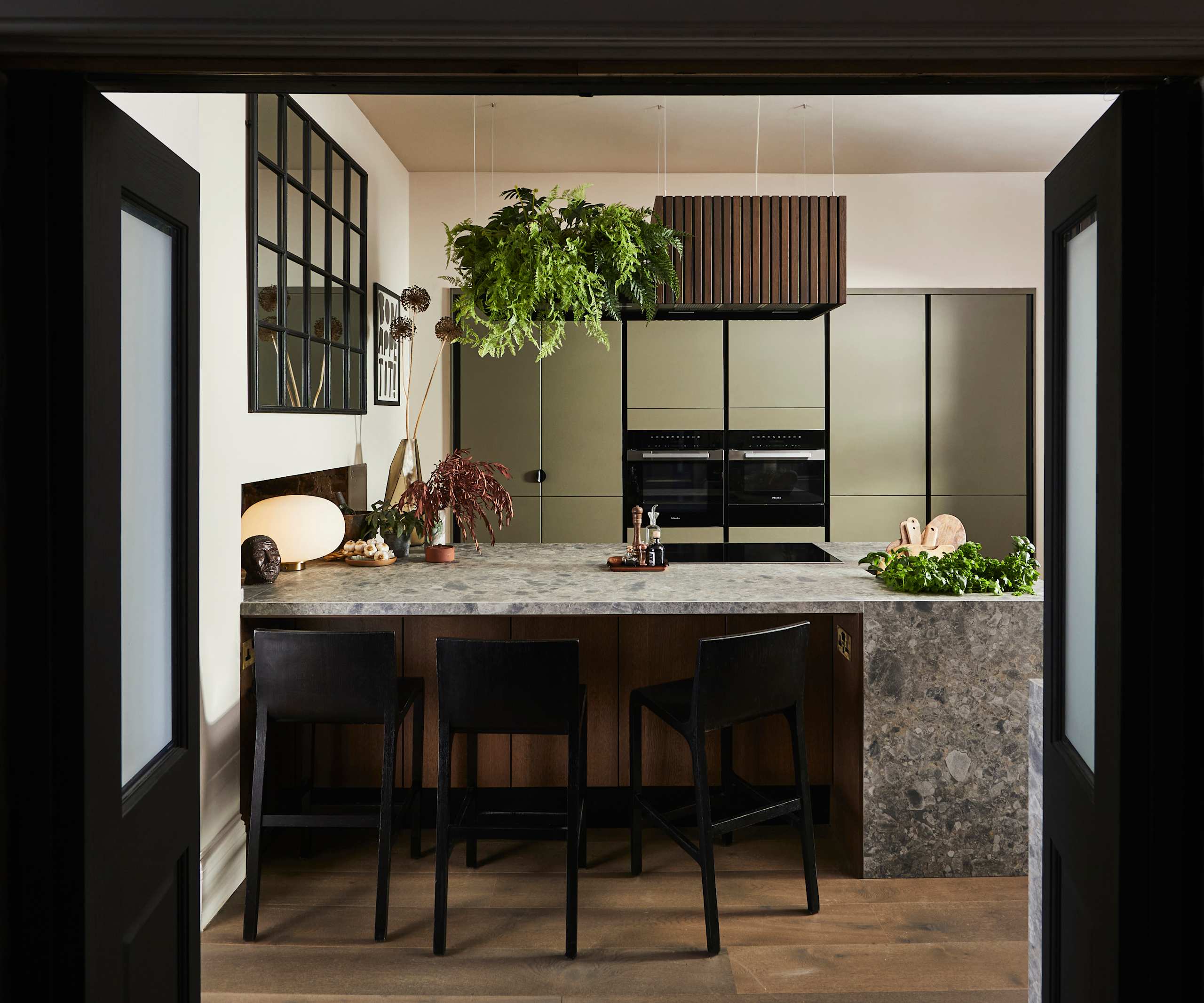 View through opened dark wood doors of the biophilic Dramatic neutral Balmoral Soho and Nordic Nature kitchen from Magnet and matching dark chairs by the bar area of the kitchen island.