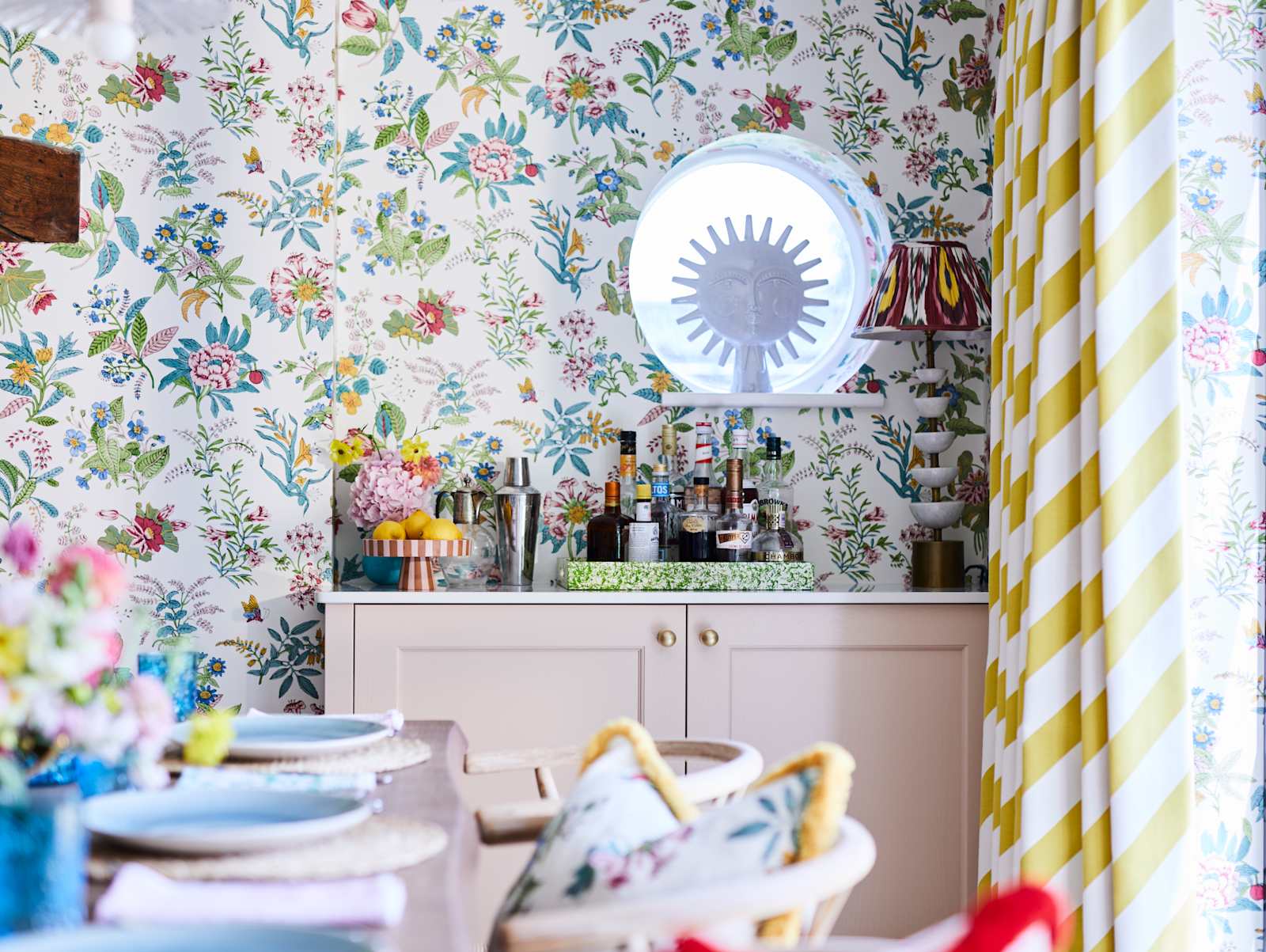 Cocktail corner in Sophie Robinsons maximalist Ludlow kitchen from Magnet, with a dining room table in foreground, floral pattern wallpaper and striped curtains, and a round window over the bar.