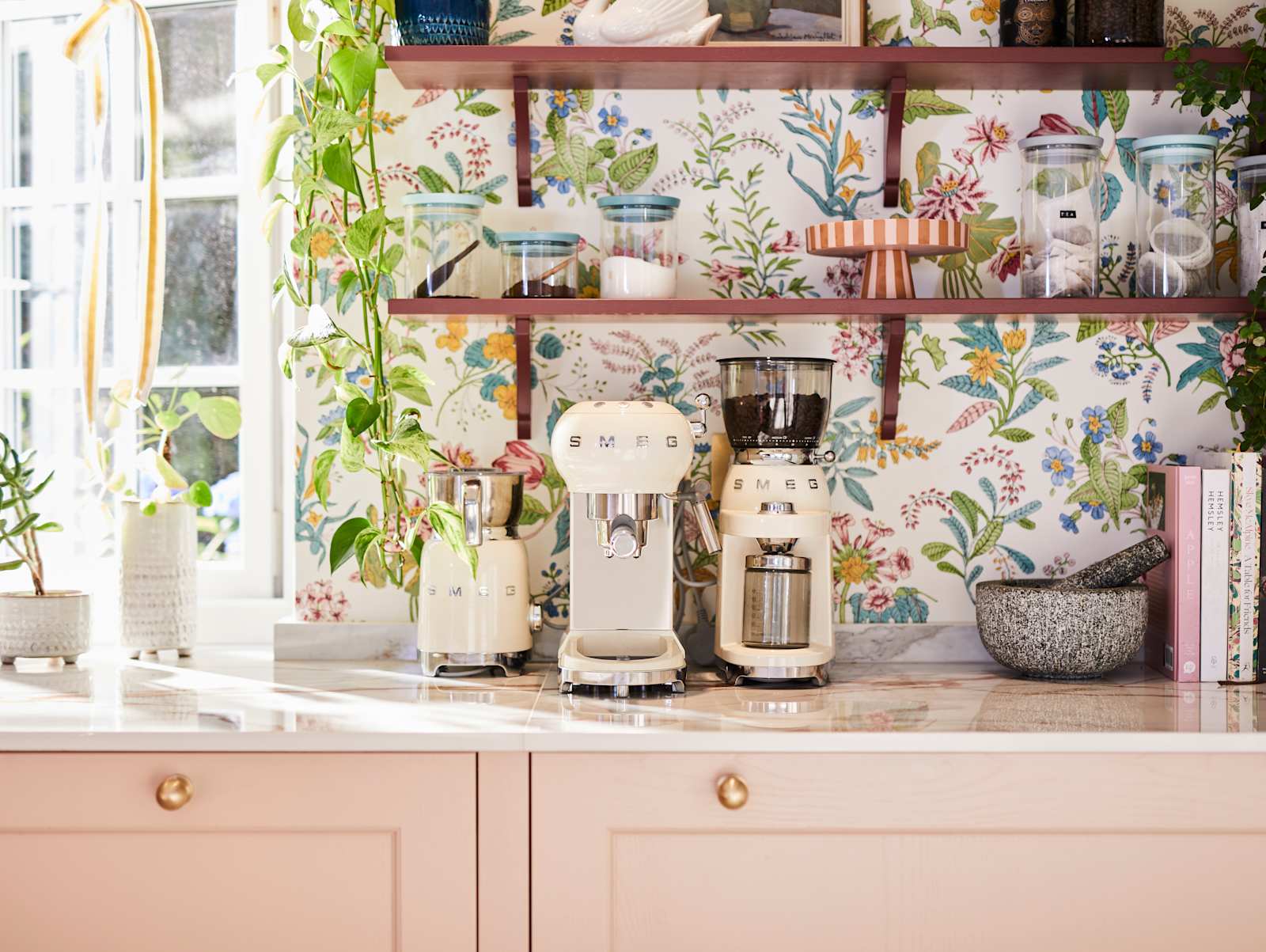 Close-up of Smeg coffee machines on counter top in the coffee area in Sophie Robinson's maximalist kitchen, with shaker style Ludlow Chalk blush cabinetry, open shelving on a wall with a floral wallpaper.