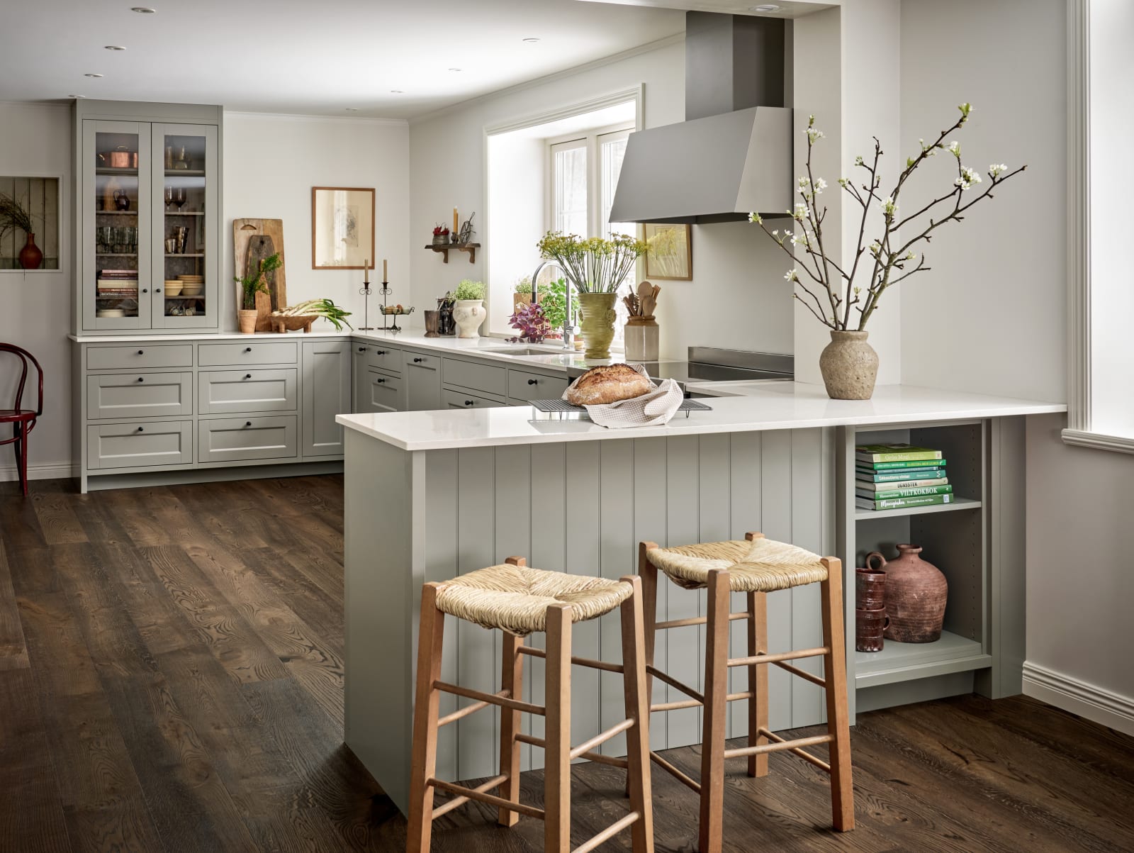 Malin Berghagens kök från Marbodal, lantliga Fagerö i kulören Frö med vit och grå marmorerad bänkskiva, med halvt fristående köksö med två barstolar och en öppen hylla för böcker och keramik.