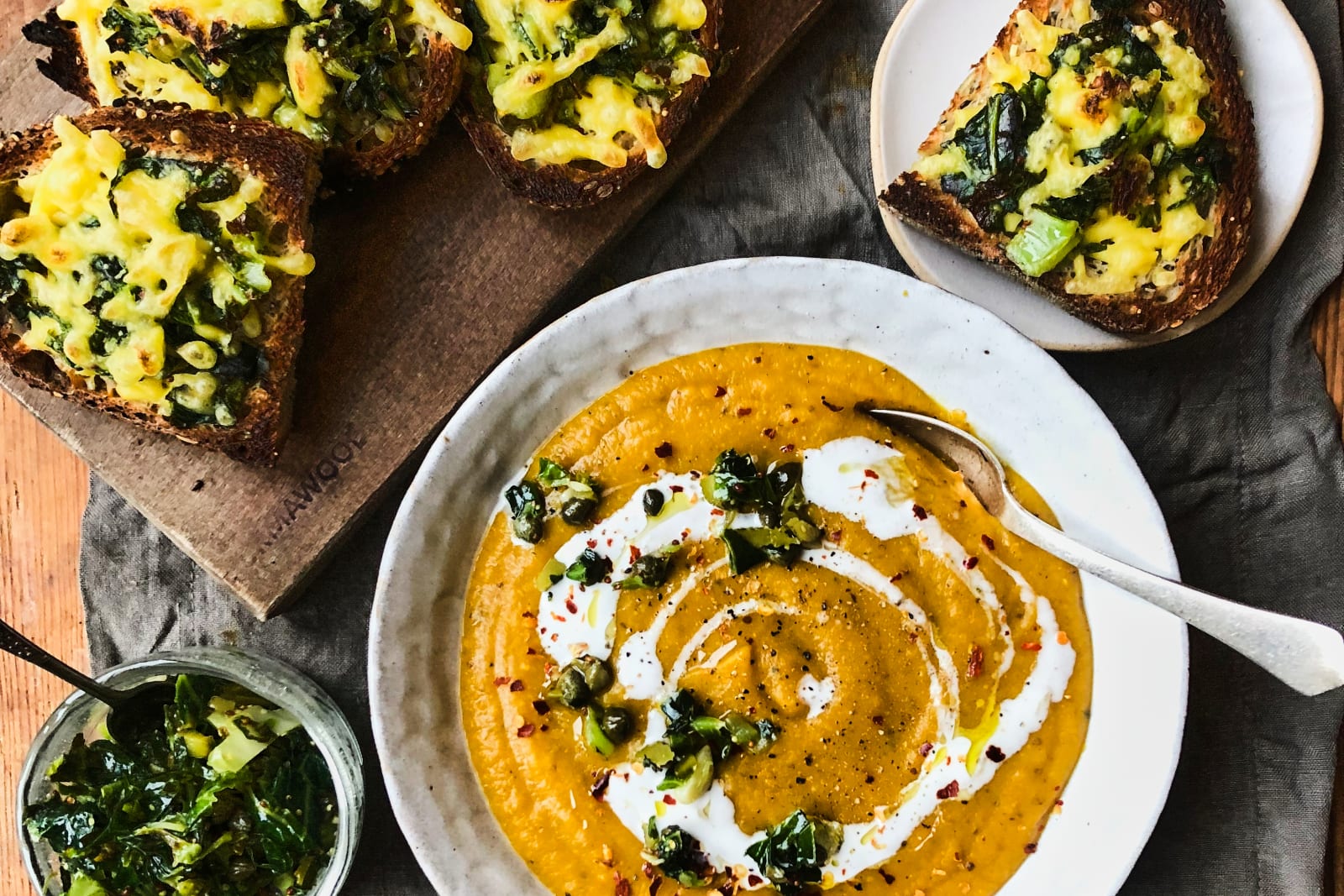 Lentil and sweet potato soup in handmade ceramics, with cheese and greens toast on a wooden cutting board, all set on a linen table cloth resting on a rustic wood table.