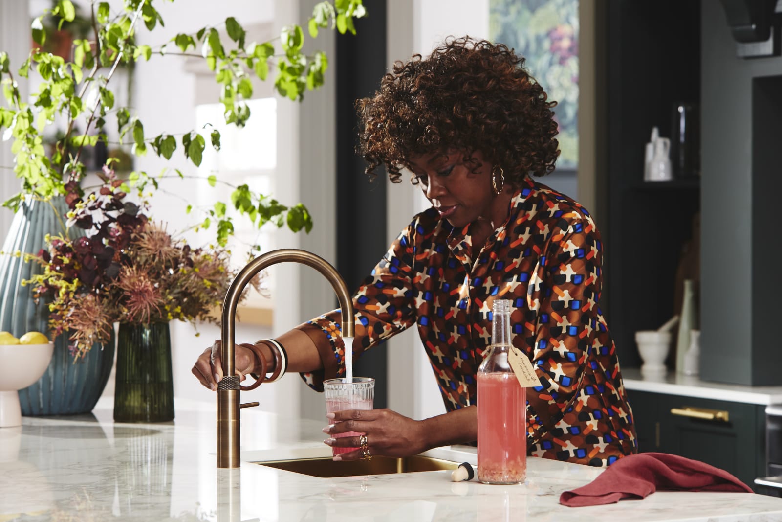 Woman puring water from tap into a glass of lemonade, with the lemonade bottle next to the sink, in a kitchen decorated with plants