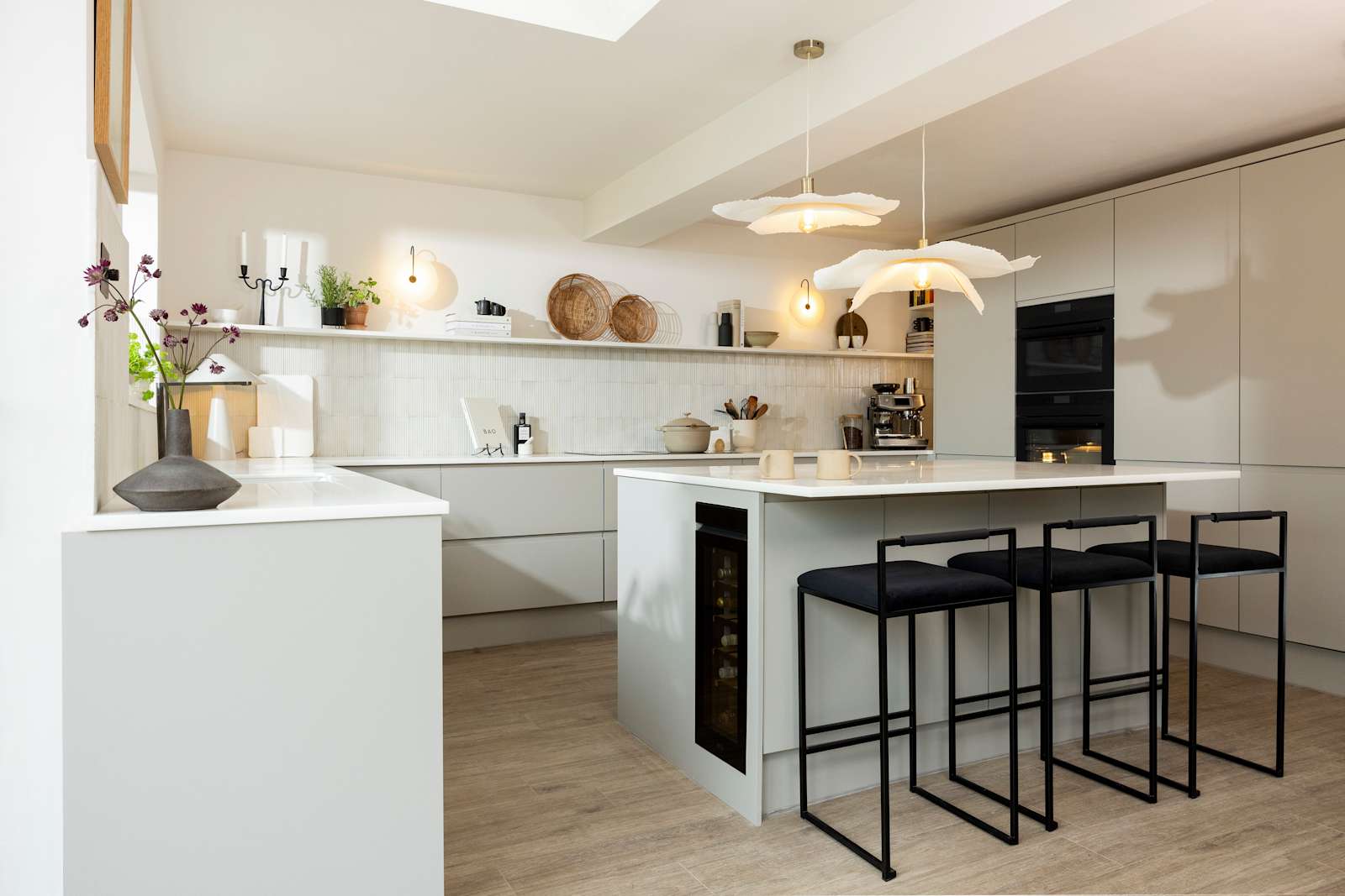 Angled view of kitchen island in Yasmin Wyatt's neutral shade Luna kitchen from Magnet with a white Corian worktop, integrated whitegoods and black metal chairs by the kitchen island bar area.