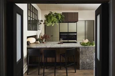 View through opened dark wood doors of the biophilic Dramatic neutral Balmoral Soho and Nordic Nature kitchen from Magnet, matching dark wood chairs by kitchen island bar seating area.