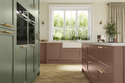 Tall cabinetry with built-in ovens and kitchen island with white worktop in Wardley kitchen from Magnet in shades Clay and Thyme, matched with brass handles and a vase of flowers on the countertop.