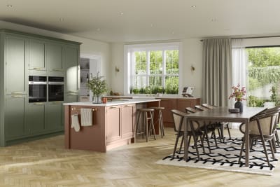 Open floorplan kitchen and dining rool with tall Wardley kitchen cabinetry from Magnet with built-in ovens and a kitchen island, in the shades Thyme and Clay, with a large wooden dining room table.