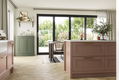 Wardley cabinetry in open floorplan kitchen and dining room with kichen island in shade Clay and a sideboard cabinet in shade Thyme, glass doors showing garden outside.