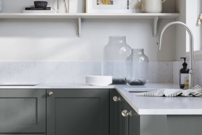 Wardley Arboretum kitchen cabinetry with steel knobs, steel tap, white marbled worktop and matching backsplash. An open shelf on the wall above cabinets with ceramics and a vase of flowers.