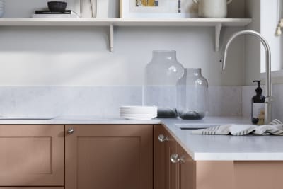 Wardley Clay kitchen cabinetry with steel knobs, steel tap, white marbled worktop and matching backsplash. An open shelf on the wall above cabinets with ceramics and a vase of flowers.