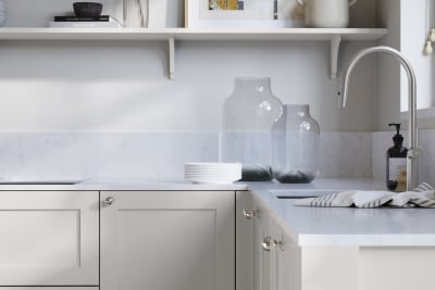 Wardley Fresh Linen kitchen cabinetry with steel knobs, steel tap, white marbled worktop and matching backsplash. An open shelf on the wall above cabinets with ceramics and a vase of flowers.
