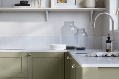 Wardley Misty Moors kitchen cabinetry with steel knobs, steel tap, white marbled worktop and matching backsplash. An open shelf on the wall above cabinets with ceramics and a vase of flowers.