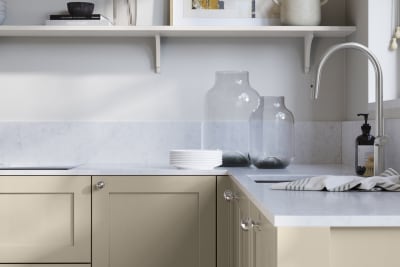 Wardley Pebble path kitchen cabinetry with steel knobs, steel tap, white marbled worktop and matching backsplash. An open shelf on the wall above cabinets with ceramics and a vase of flowers.