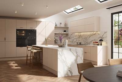 Fluted front kitchen Reed from Magnet in light warm neutral shade Shell, with a marbled splashback, worktop and decorative side on kitchen island with barstools and black handles, herringbone wood flooring.