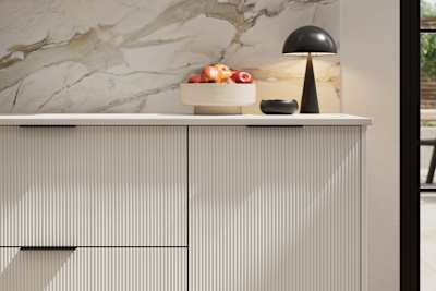Fluted front kitchen Reed from Magnet in light shade Fresh Linen, with a marbled splashback and black handles, herringbone wood flooring.