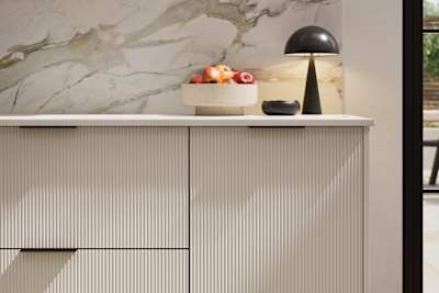 Fluted front kitchen Reed from Magnet in light neutral shade Limestone, with a marbled splashback and black handles, herringbone wood flooring.