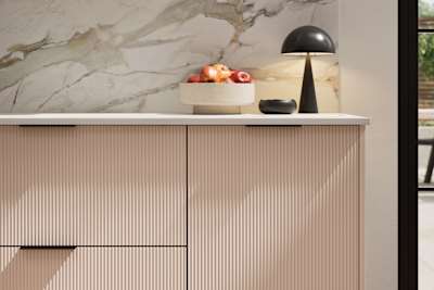 Fluted front kitchen Reed from Magnet in light neutral pink shade Chalk Blush, with a marbled splashback and black handles, herringbone wood flooring.