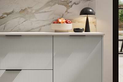Fluted front kitchen Reed from Magnet in light neutral shade Washed cotton, with a marbled splashback and black handles, herringbone wood flooring.