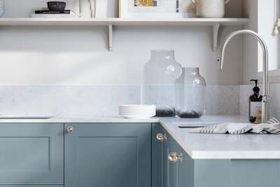 Ludlow, a traditional Shaker-style premium feel kitchen range with a visible woodgrain on its panelled doors here shown in the shade Portland Blue styled with steel knobs, steel tap, a white marbled worktop with matching splashback.