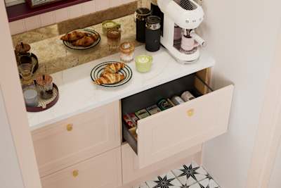 Opened drawer of teas in painted wood grain Shaker style Bewdley cabinets in light pink shade Chalk Blush, with marbled worktop, brass knobs and Smeg coffee machine on countertop.