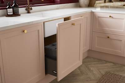 Opened sink drawer in painted wood grain Shaker style Bewdley kitchen with cabinets in light pink shade Chalk Blush, marbled worktop and brass knobs.