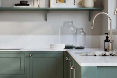 Bewdley Eucalyptus Green painted wood grain Shaker kitchen cabinetry with steel knobs, steel tap, white marbled worktop and matching backsplash, an open shelf on the wall above cabinets with ceramics and a vase of flowers.