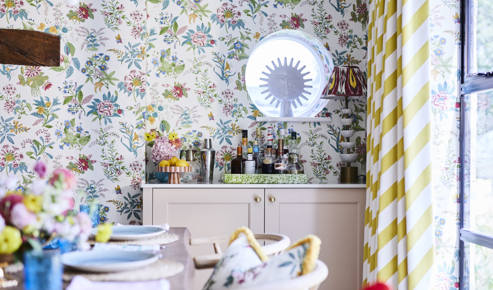 Cocktail corner in Sophie Robinsons maximalist Ludlow kitchen from Magnet, with a dining room table in foreground, floral pattern wallpaper and striped curtains, and a round window over the bar.