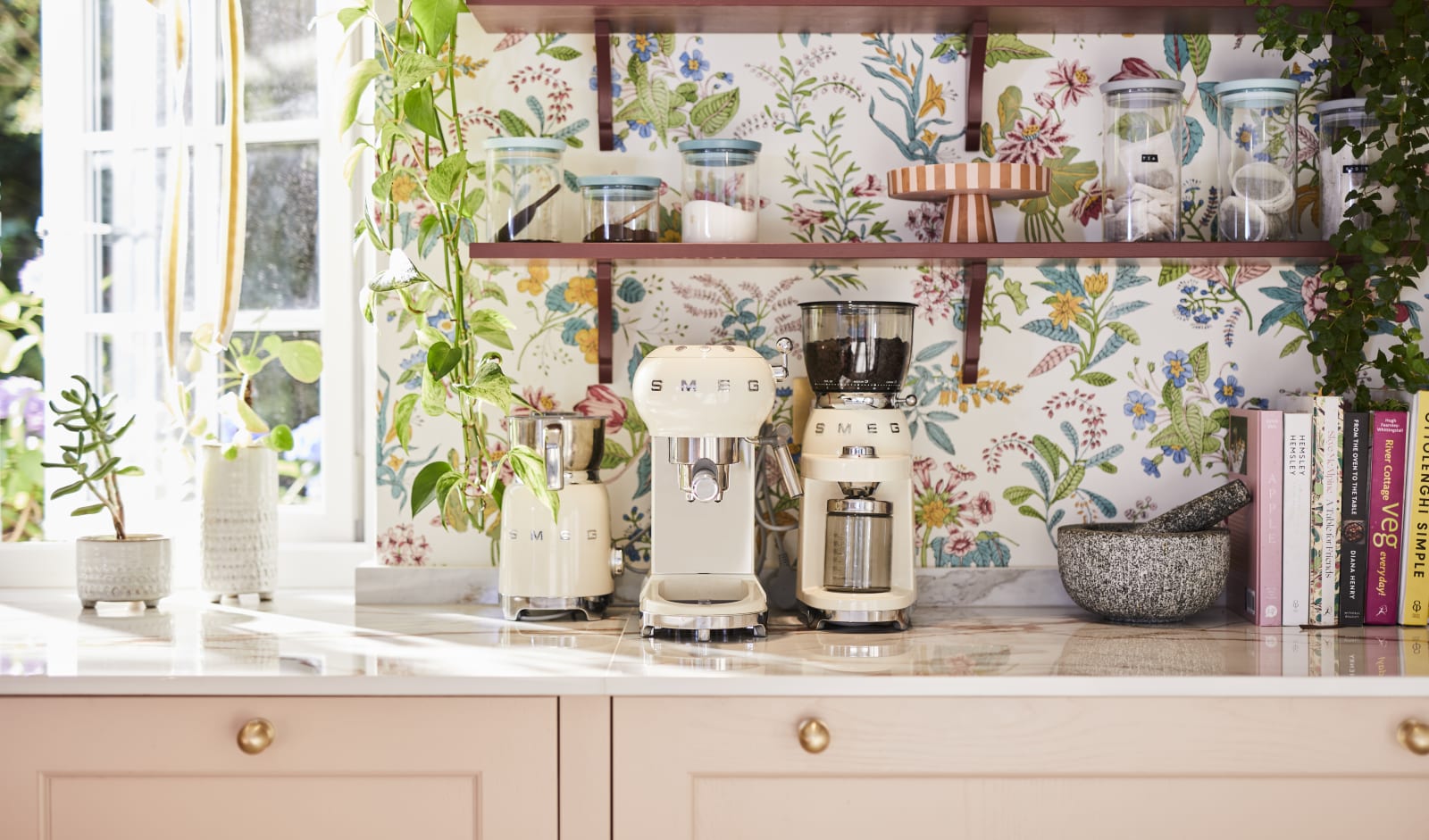 Close-up of Smeg coffee machines on counter top in the coffee area in Sophie Robinson's maximalist kitchen, with shaker style Ludlow Chalk blush cabinetry, open shelving on a wall with a floral wallpaper.