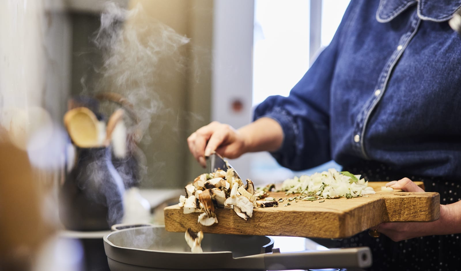 Close-up of Jen pouring mushrooms and vegetables from a thick wooden cuttingboard into a pan on the stove, with stem rising from the pan