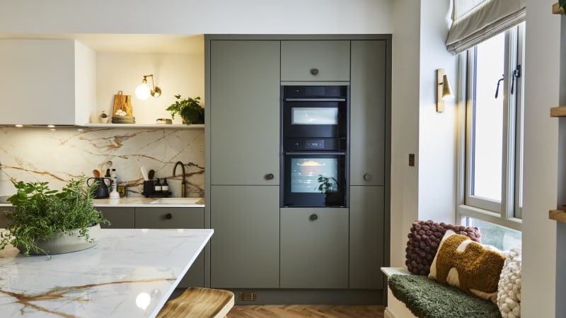Built-in ovens in tall slab Soho Balmoral cabinets, a bench with earth tone pillows and a kitchen island with a wooden barstool and a plant