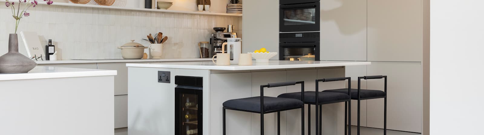 Diagnonal view of kitchen island corner in Yasmin Wyatt's open, light and airy neutral shade handleless Luna kitchen from Magnet with a white Corian worktop in Onyx, inegrated white goods and a bar area of kitchen island with black barstools.