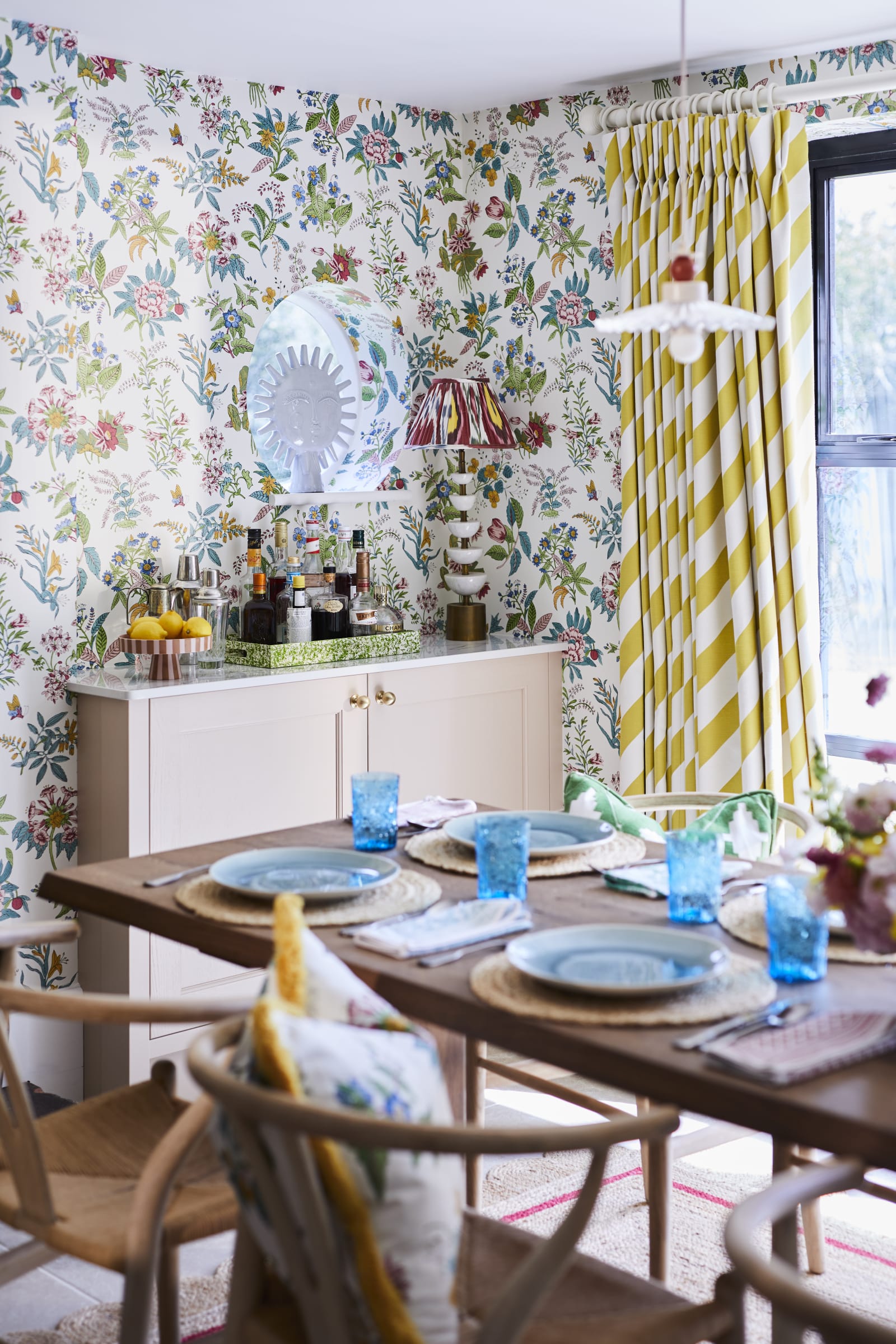 Cocktail corner in Sophie Robinsons maximalist Ludlow kitchen from Magnet, with a dining room table in foreground, floral pattern wallpaper and striped curtains.