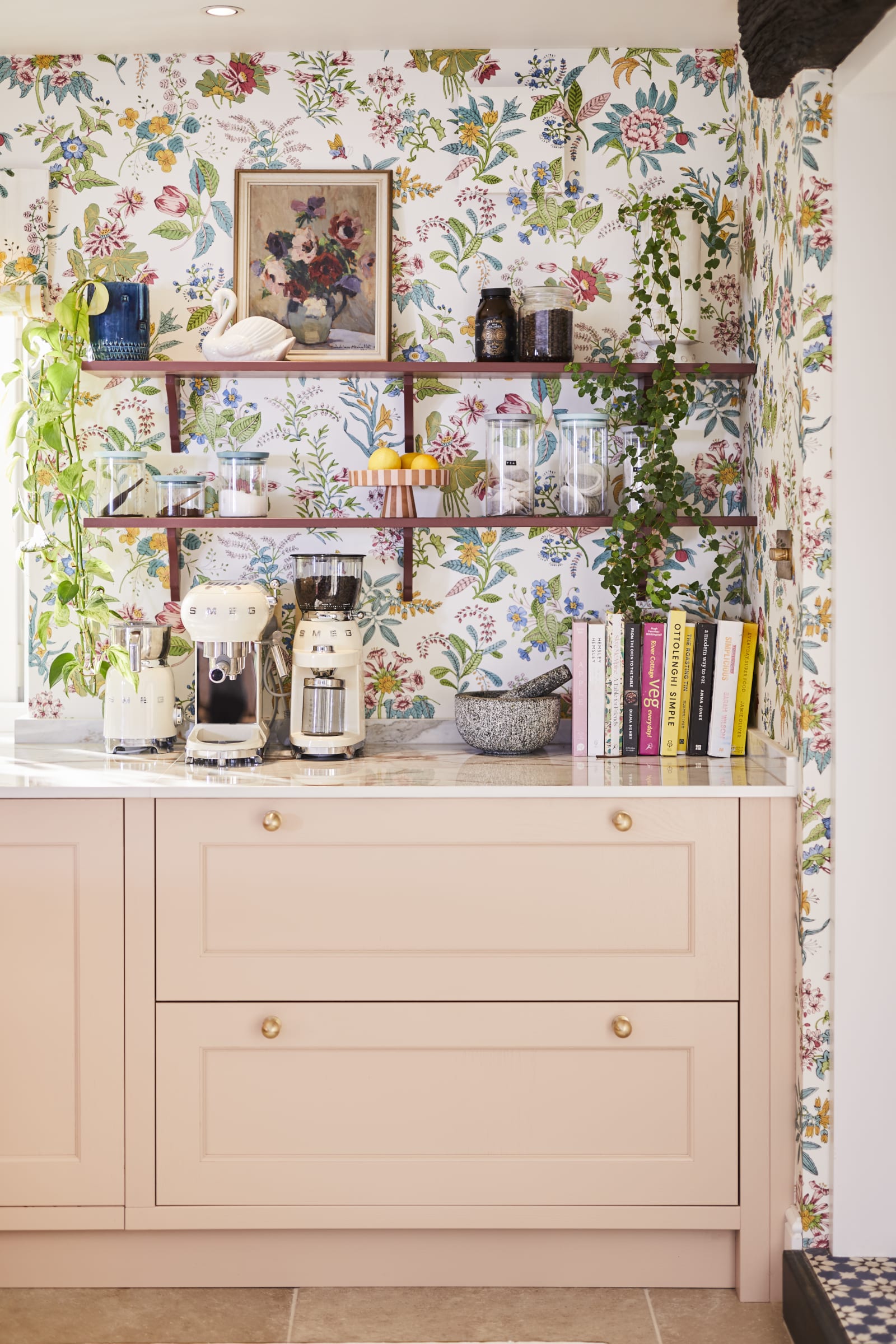 Coffee area in Sophie Robinson's maximalist kitchen, with shaker style Ludlow Chalk blush cabinetry, open shelving on a wall with a floral wallpaper and Smeg coffee machines on countertop.