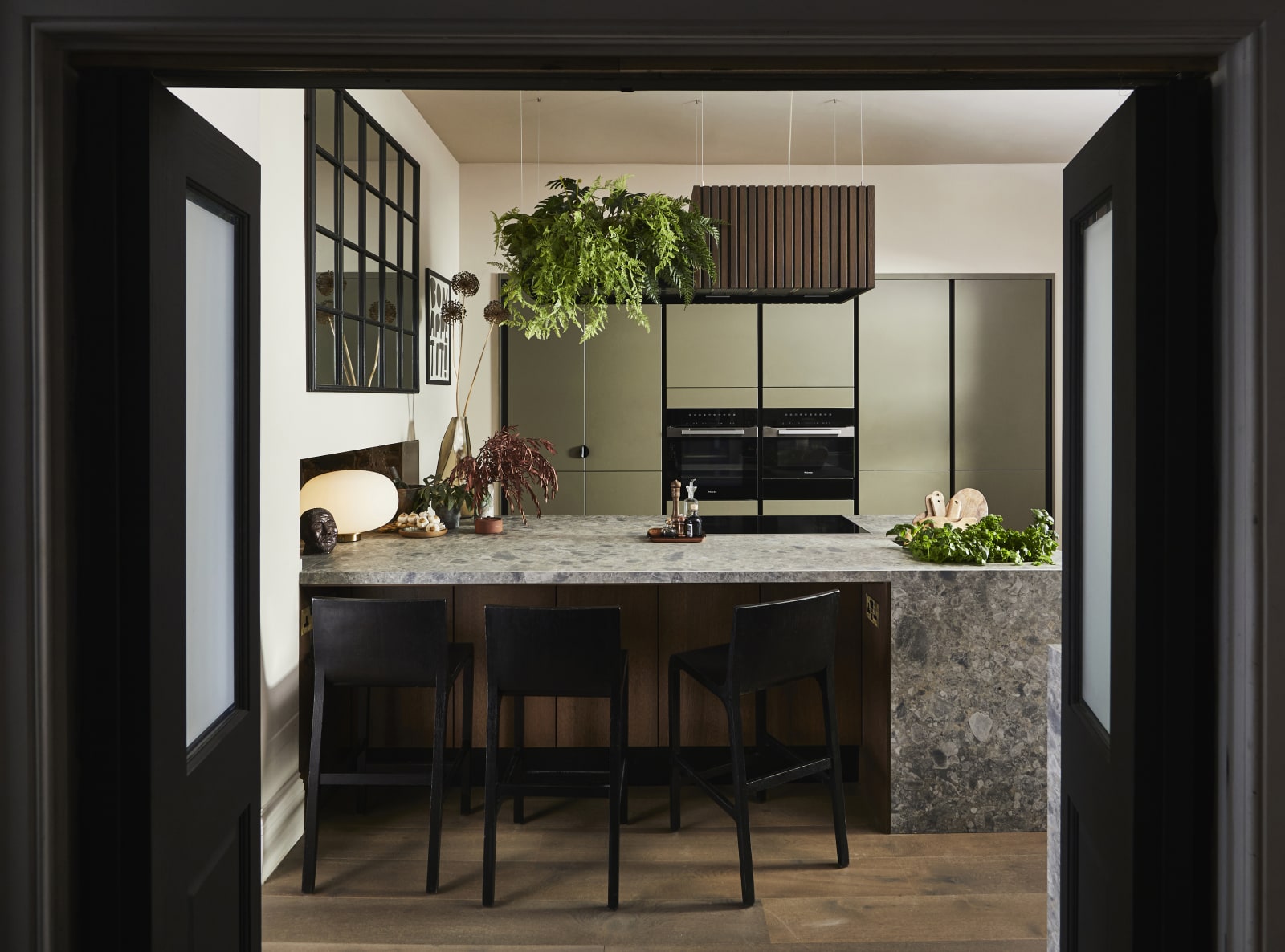 View through opened dark wood doors of the biophilic Dramatic neutral Balmoral Soho and Nordic Nature kitchen from Magnet and matching dark chairs by the bar area of the kitchen island.