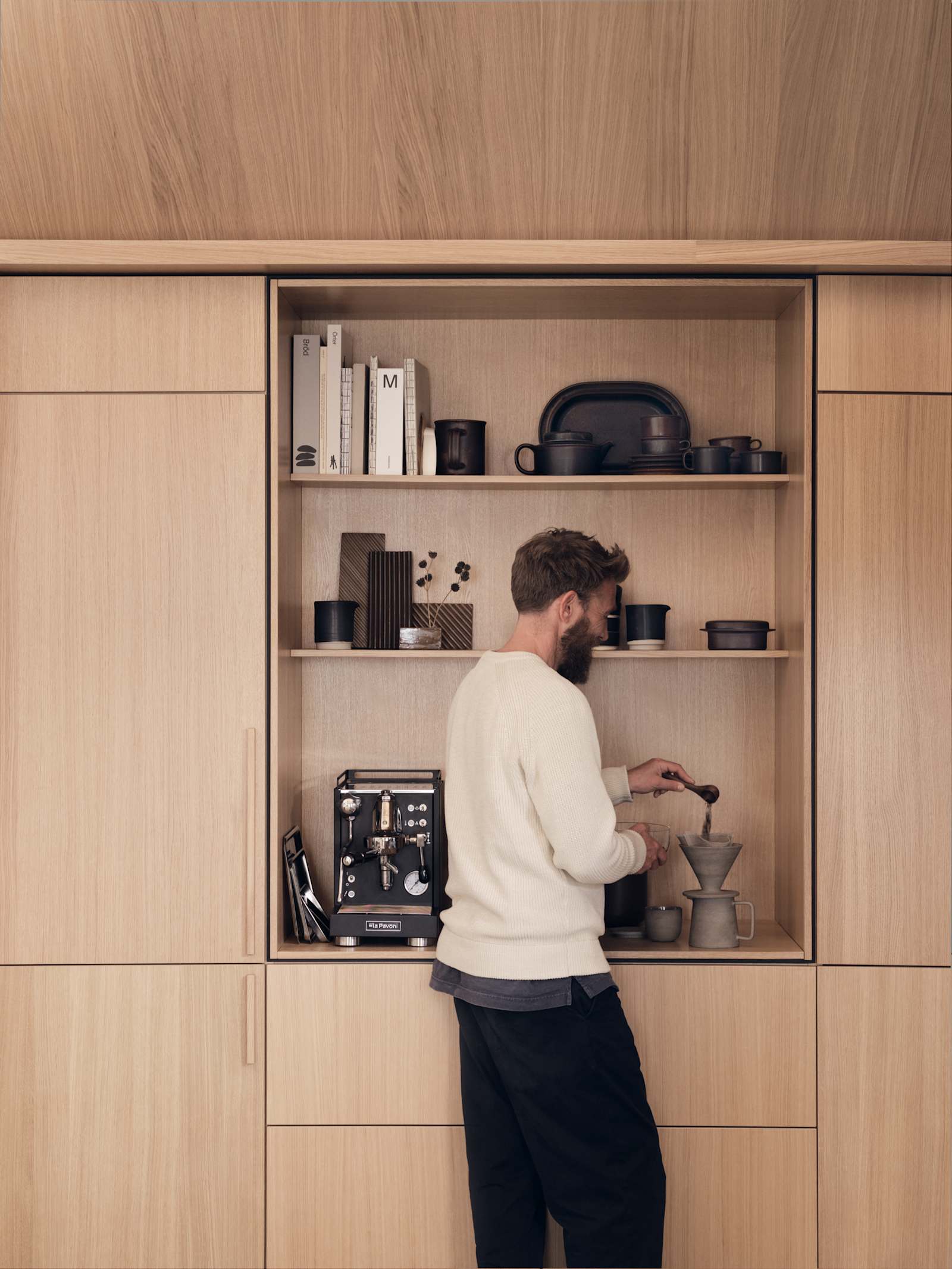 En mand måler kaffe op i Forever Oak Natur Eg-køkkenet fra INVITA i egetræsfiner med lodret træåring. Højskabe og et åbent skab med plads til kaffemaskine, indrammet med en skygge, der skaber en arkitektonisk premium følelse.