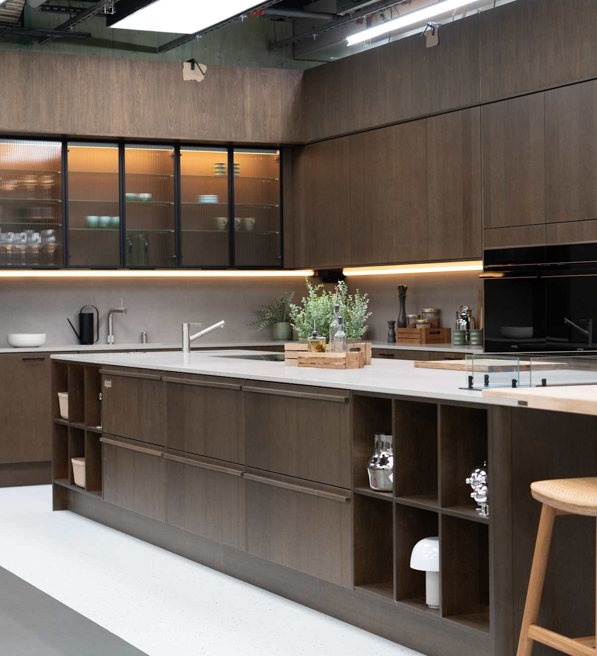 Kjøkken i modell Favn fra Sigdal i lekker eikefinér, som er beiset i fargen Nøtt, kjøkkenøya og vitrinedører med bølget glass og sorte aluminiumsrammer i God Morgen Norge studion sin