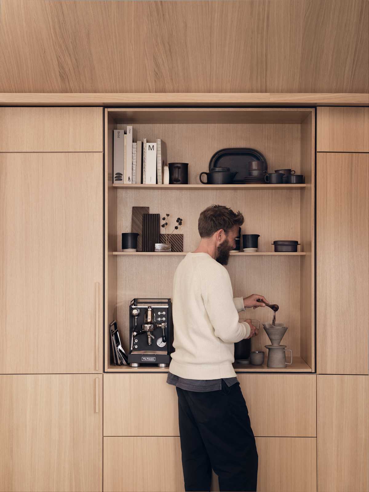En mann måler opp kaffe ieikekjøkkenet Favn Natur fra Sigdal i eikefiner med vertikal trestruktur. Høyskap og et åpent skap med plass til kaffemaskin, innrammet med en skyggelegging som skaper en arkitektonisk premiumfølelse.