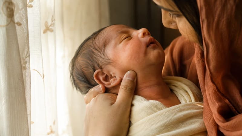 A tender, warm-lit editorial photograph of a South Asian mother gently assessing her newborn baby's cheek and neck skin under soft window daylight, illustrating tactile assessment of hyperpigmented and violaceous skin inflammation.