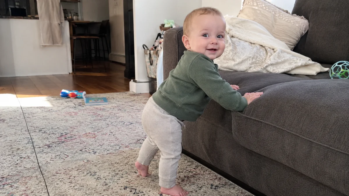 An 11-month-old baby pulling up to stand and cruising along a sofa, a late first-year motor milestone that usually comes just before walking.