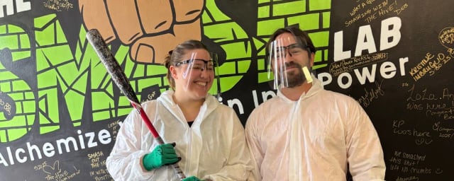 Two people in white protective suits and green gloves posing with baseball bats before a mural — The Smash Lab LLC, from its own website