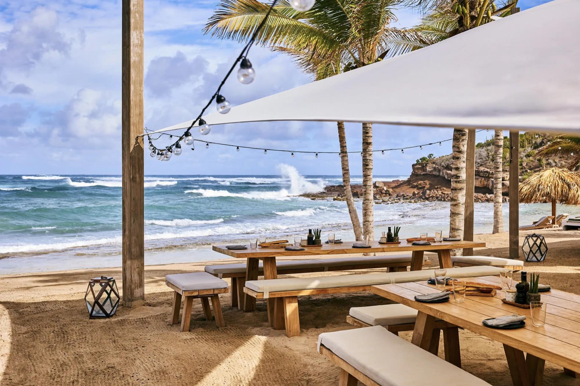 Table set on the Guanahani beach under fairy lights, wooden benches and view of the bay