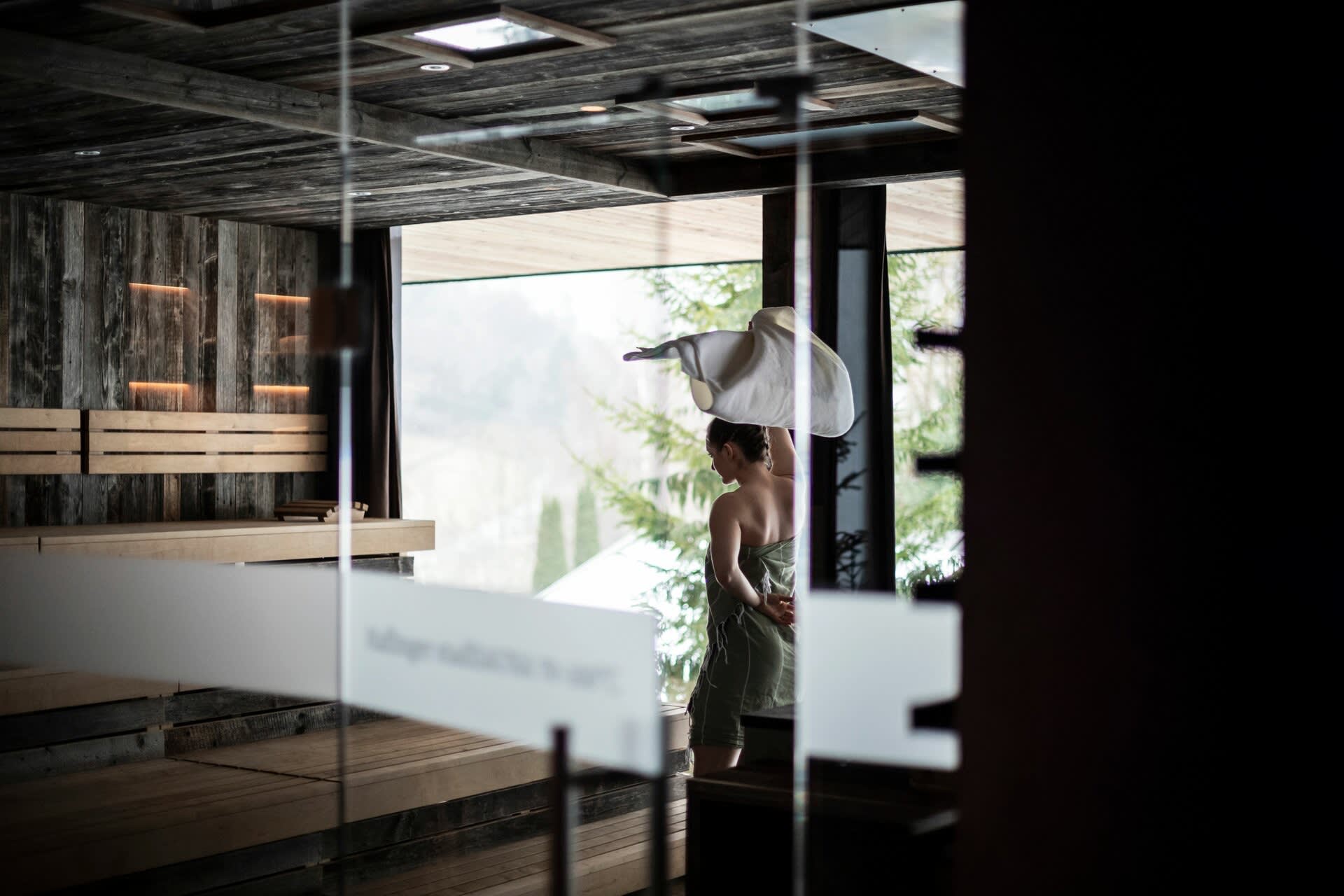 Panoramic sauna at the Viktoria Alpine Hideaway, floor-to-ceiling window overlooking the forest and a woman in a bathrobe