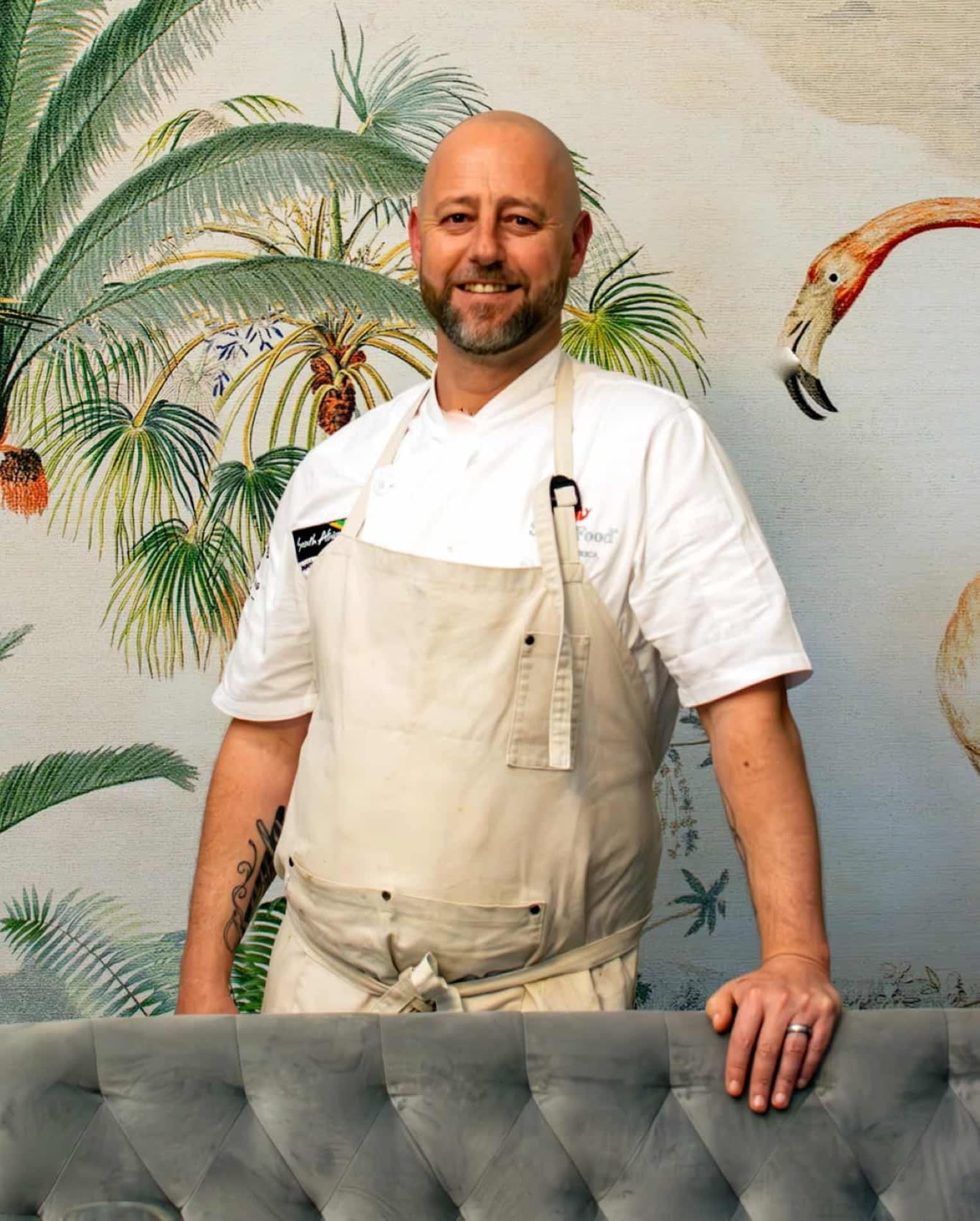 Le chef exécutif Gregory Henderson du Palm House au Cap, en veste blanche devant une fresque de palmiers et de flamants roses